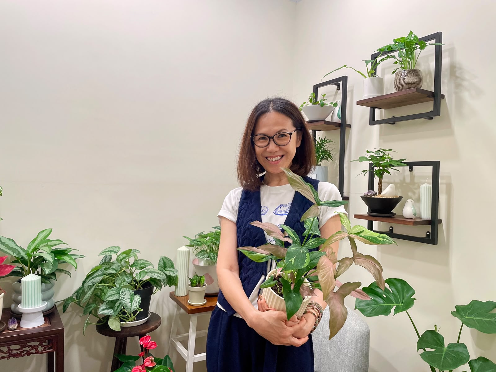 Anita stands indoors behind a large potted houseplant with green and silver variegated leaves. The background shows a cream-colored wall with black metal shelving holding other small plants.